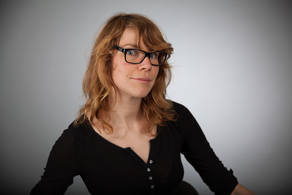 Porträtieren von Adrienn  Adrienn mit RotBlonden Haaren, Brille mit Schwarzen Gestell und Schwarzen Oberteil steht im Fotostudio vor einer Grauenfotowand.